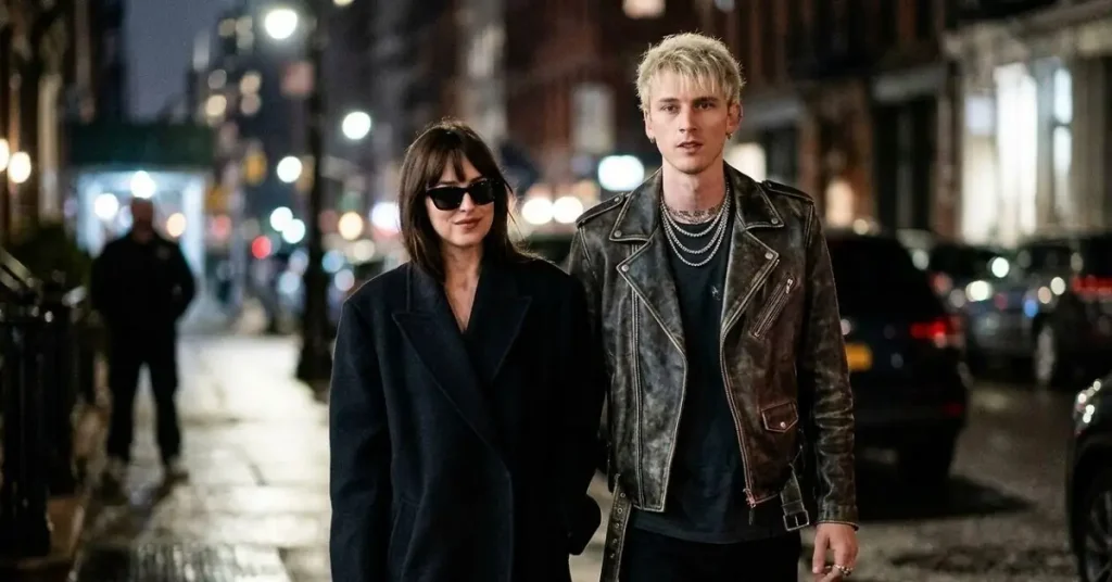 Dakota Johnson wearing a black oversized coat and sunglasses walking side-by-side at night with Machine Gun Kelly in a distressed leather jacket and silver chains.
