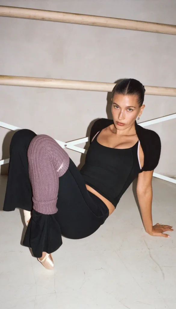 Hailey Bieber posing in a ballet studio wearing a black top and purple leg warmers
