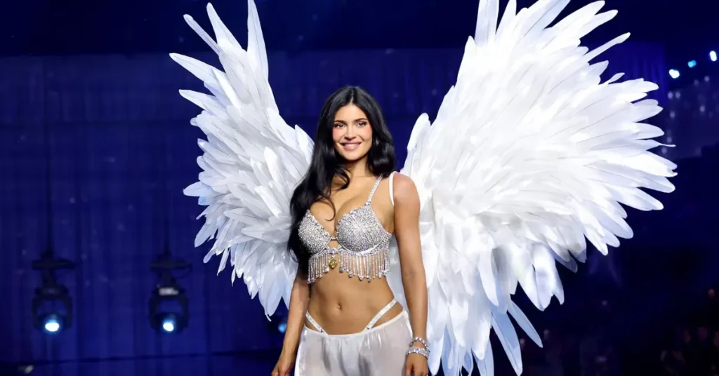 Kylie Jenner showcases an elaborate jeweled top with oversized white wings on a blue-lit fashion runway.