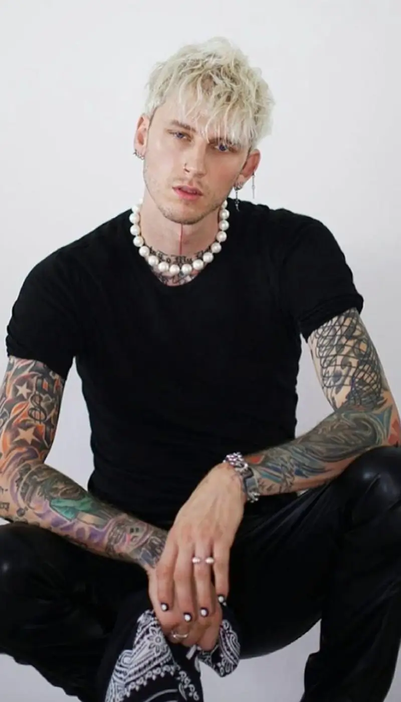 Machine Gun Kelly crouching with tattoos on his arms, wearing a fitted black t-shirt, pearl necklace, black leather pants, and painted nails.