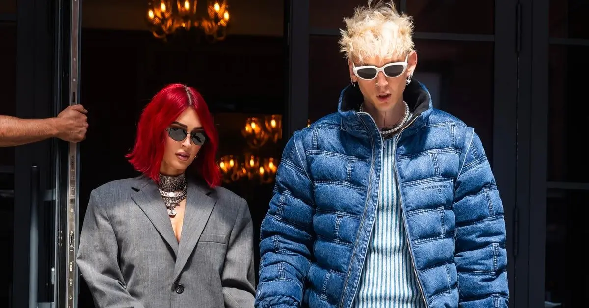 Megan Fox with bright red hair wearing a grey oversized blazer dress and snakeskin boots holding hands with Machine Gun Kelly in a blue denim puffer jacket and cargo jeans.